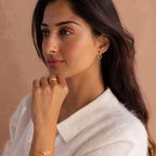 A woman with long dark hair, wearing a white top and elegant Dainty Duo Pave Huggies, featuring interlocking circles, gazes thoughtfully to the side.
