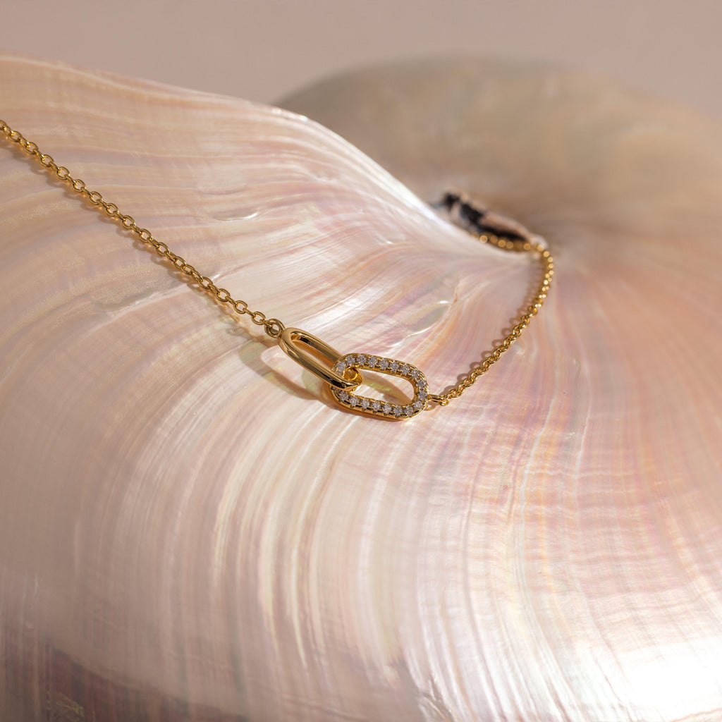 Gold chain bracelet displayed on a pearlescent seashell, featuring two interlocking oval links at the center, one polished and the other encrusted with small clear stones.