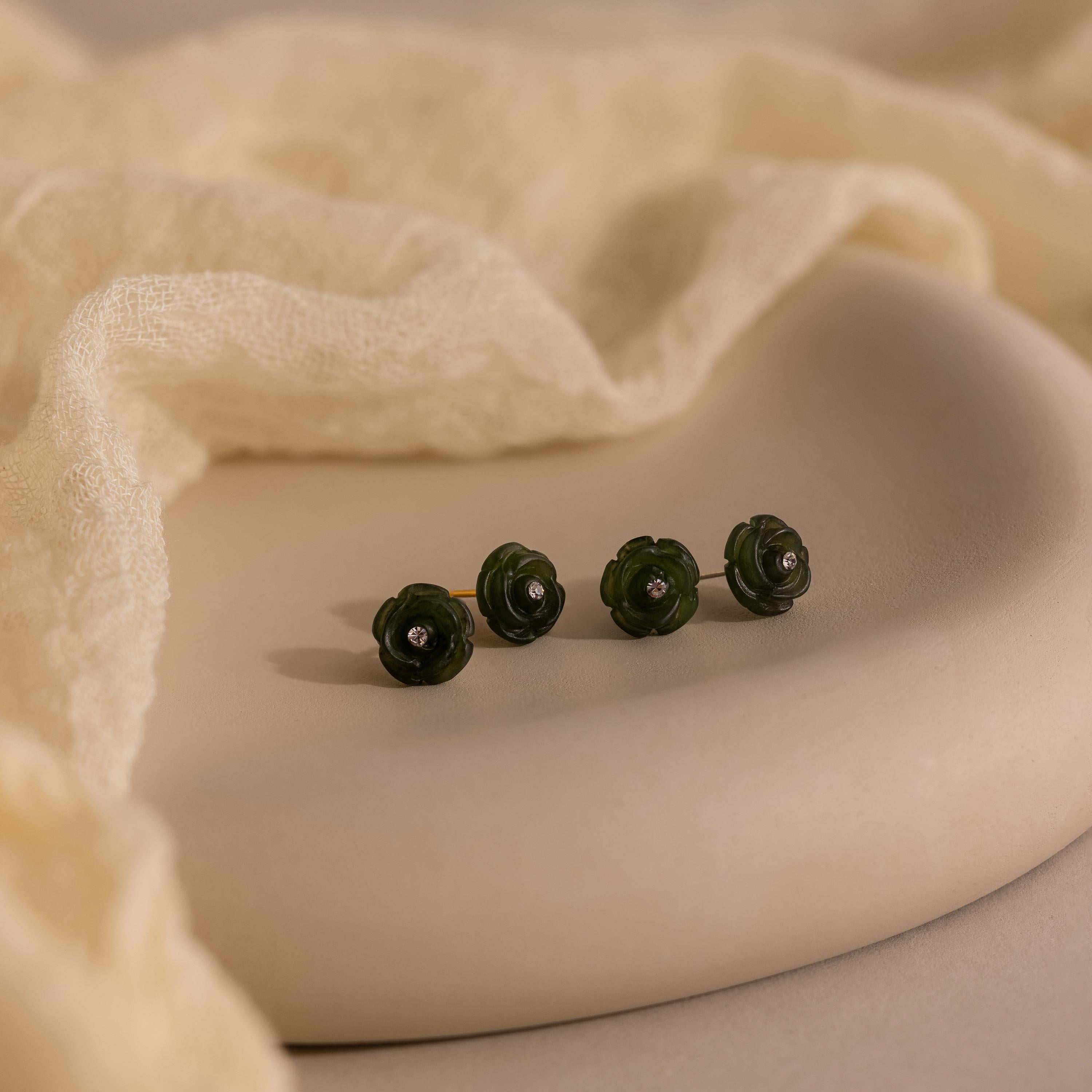Four green Jade Rose Studs, each shaped like a delicate flower with a diamond center, are displayed on a soft beige surface with fabric in the background.