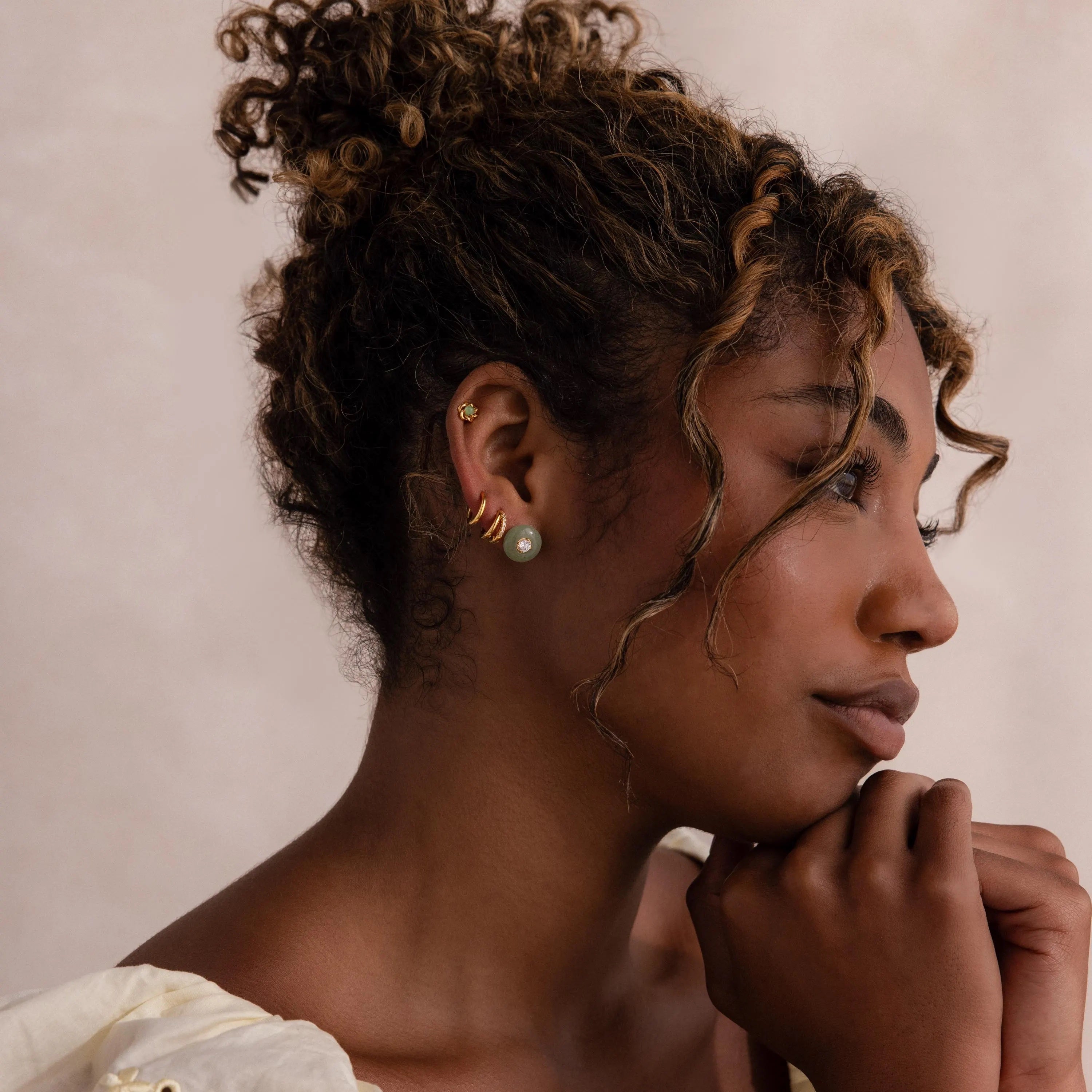 Far-view of model wearing a gold flower-shaped jade stud earring in her cartilage, stacked with a round jade earring, and multiple gold huggie hoops.