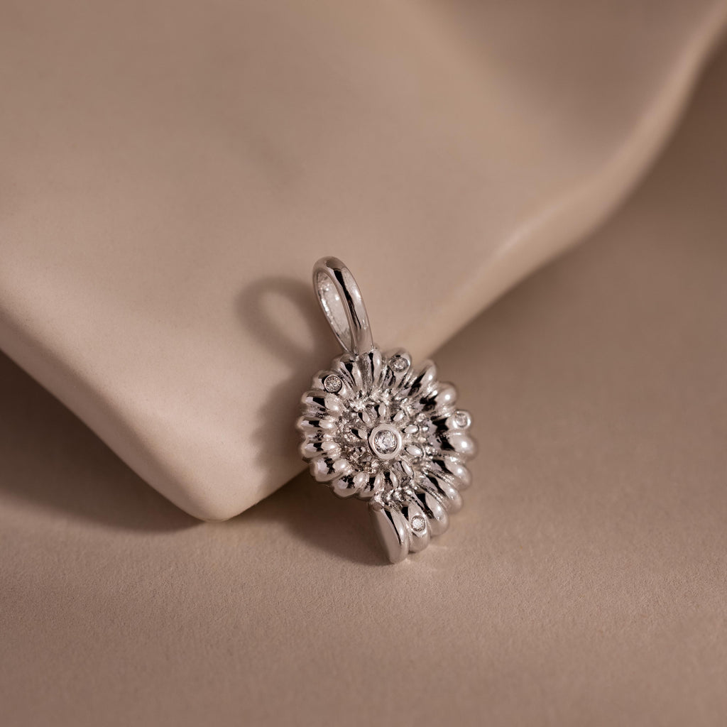 Silver shell-inspired charm pendant with a textured circular design and gemstone accents, displayed on a soft neutral fabric background.