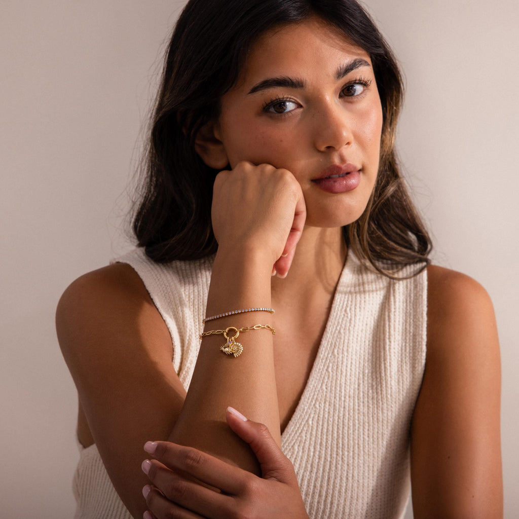 Woman wearing a gold chain bracelet with a shel inspired charm layered alongside a delicate gemstone tennis bracelet, styled with a sleeveless cream knit top.