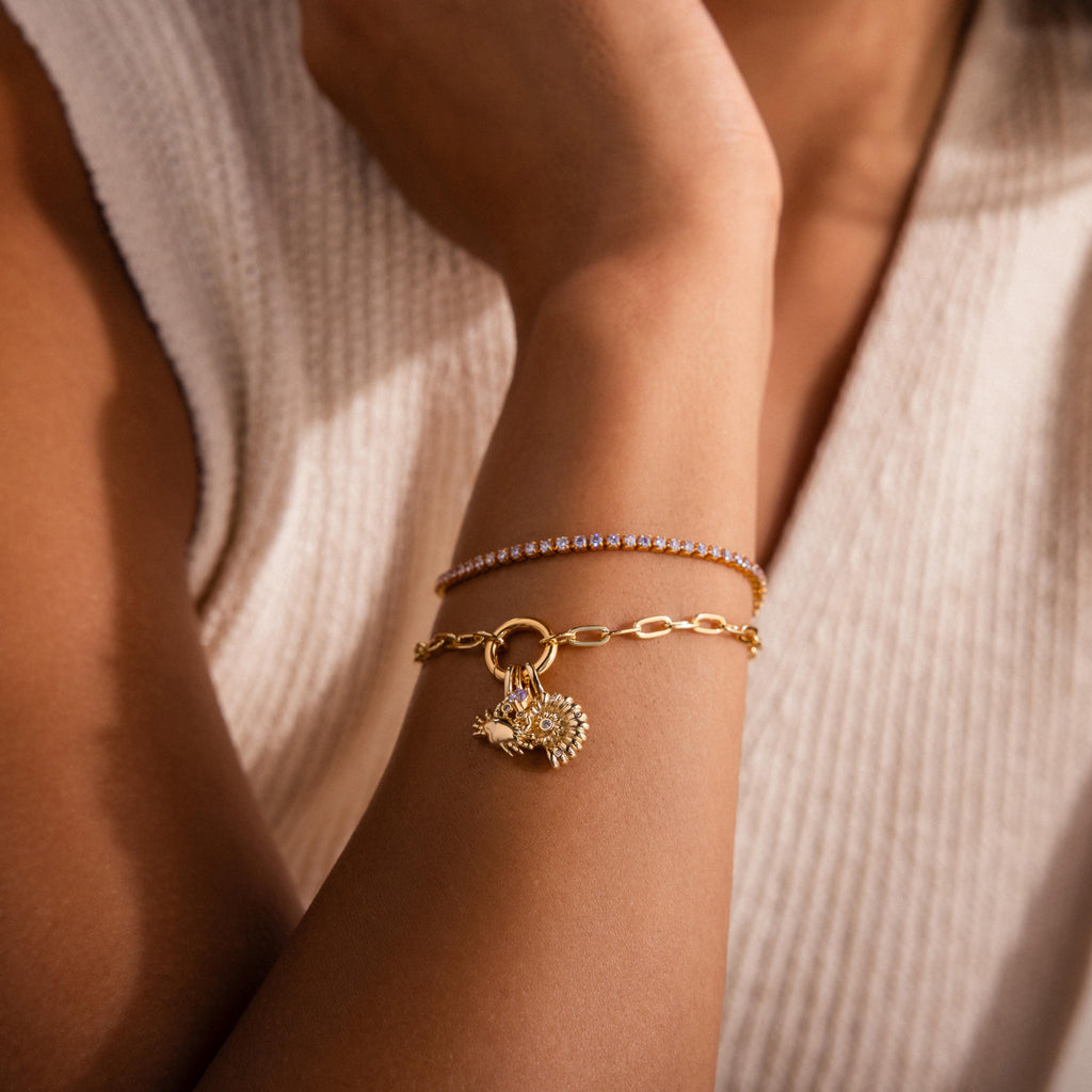Gold chain bracelet with shell inspired charm and crab charm, layered with a delicate gemstone tennis bracelet, styled on the wrist with a cream knit top.