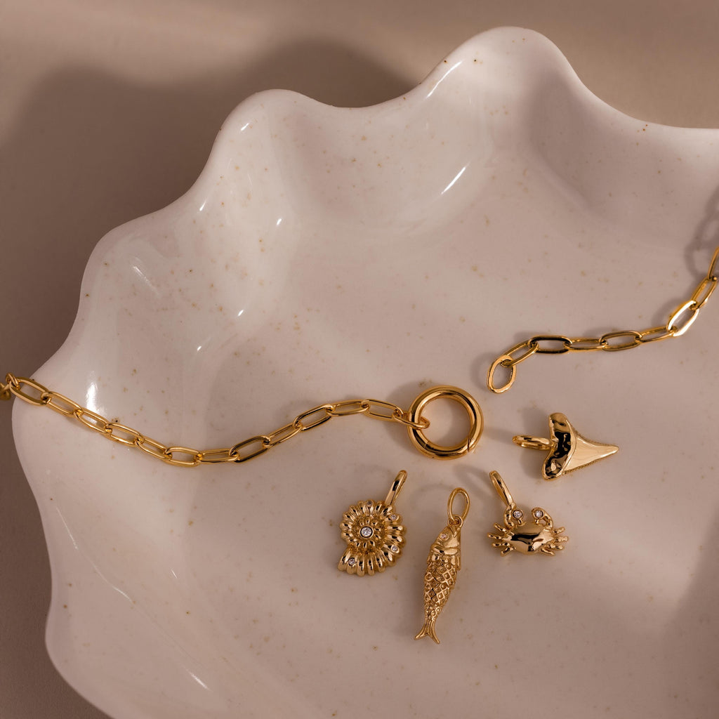 The Crab Charm features on a gold chain with four ocean-themed charms, elegantly displayed on a wavy ceramic dish—perfect beach jewelry to complement your seaside-inspired style.