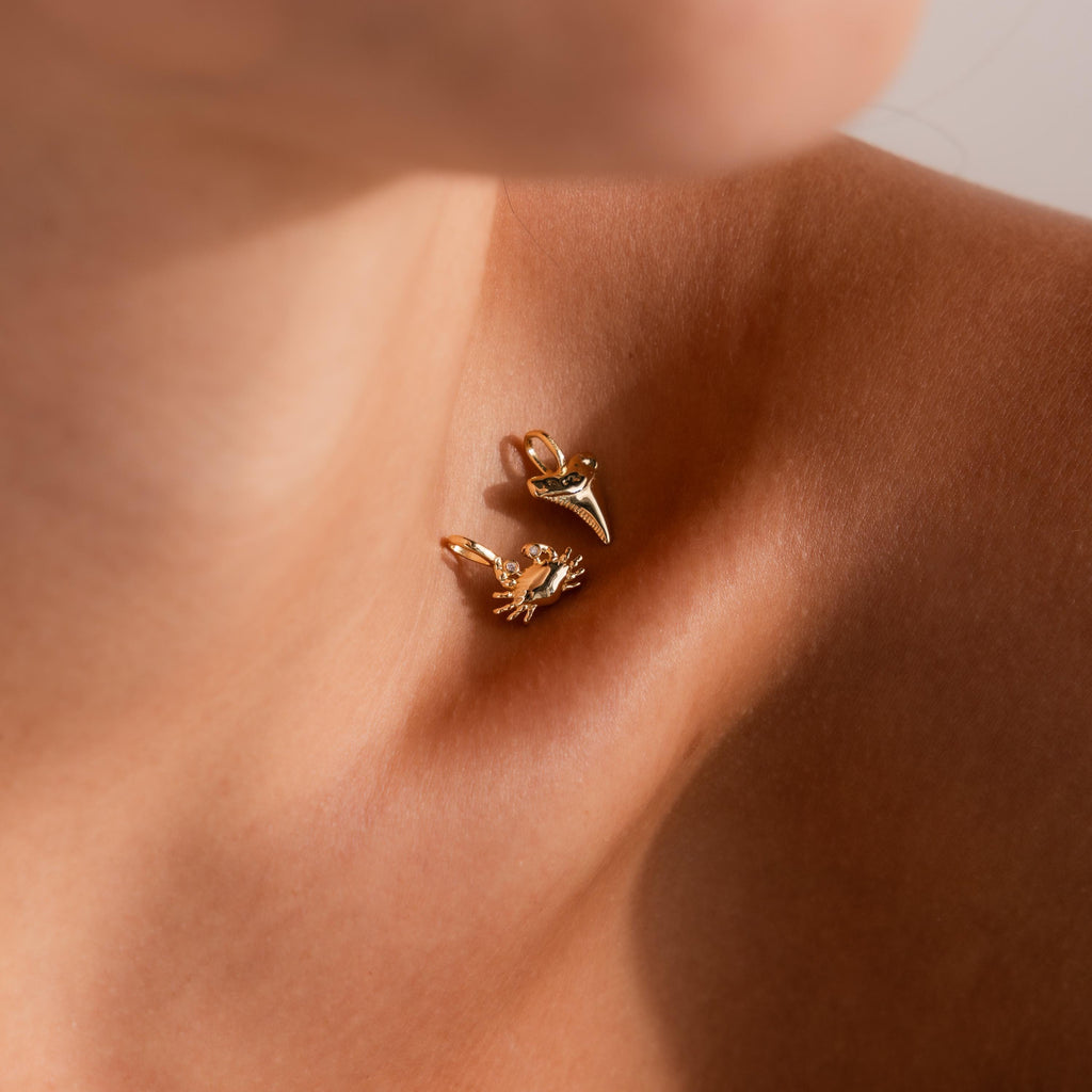 Close-up of a woman's collarbone adorned with two gold charms, a detailed crab charm and a polished shark tooth charm, both catching the light against bare skin.