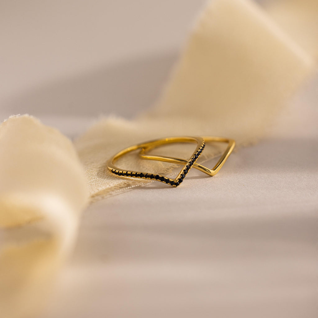 Gold chevron stacking rings, one with black stones and one plain band, displayed on soft beige fabric.