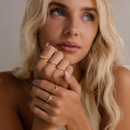 Woman with blonde hair wearing multiple gold diamond rings, including heart curve cluster designs, styled on both hands against a neutral background.