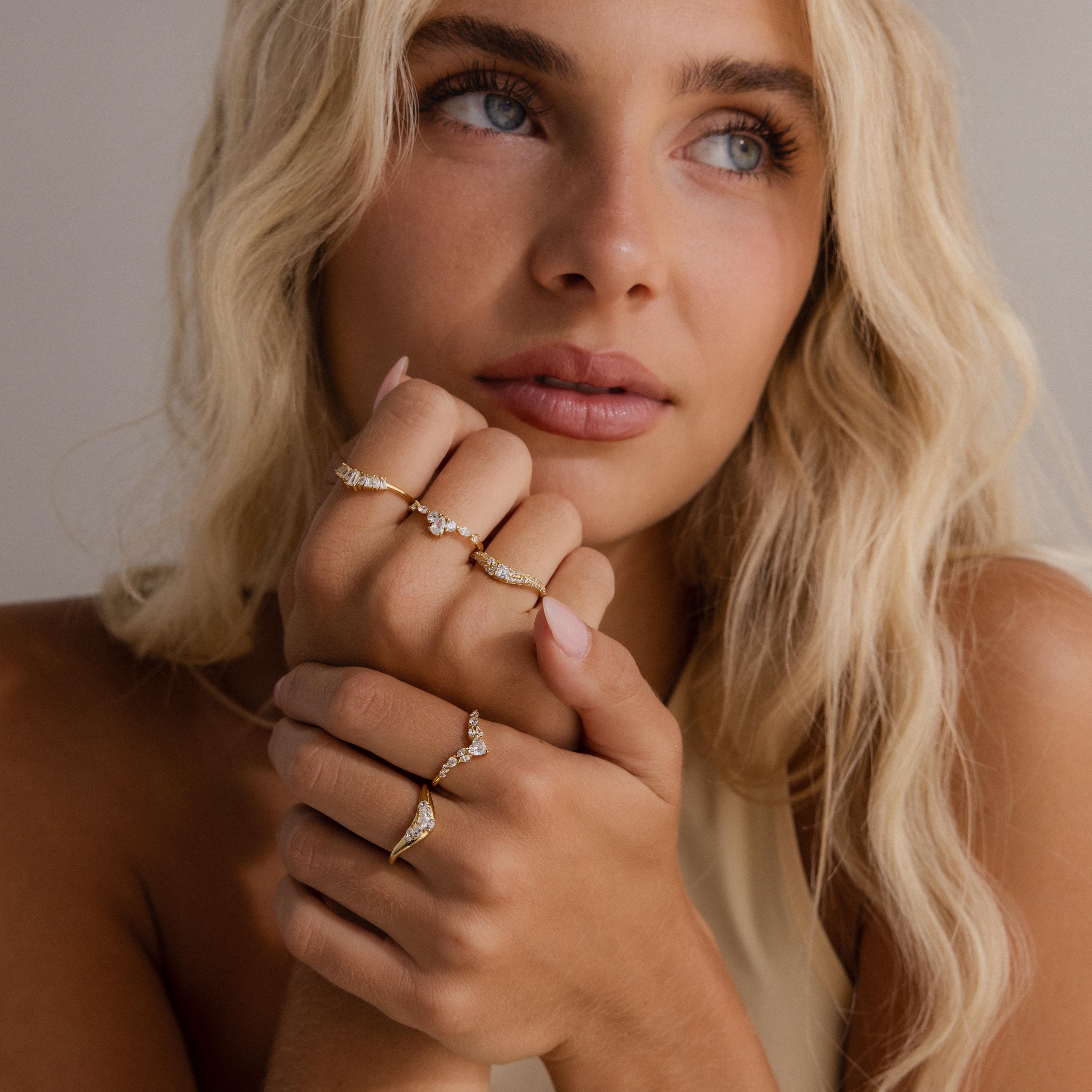 Woman with blonde hair wearing multiple gold diamond rings, including heart curve cluster designs, styled on both hands against a neutral background.
