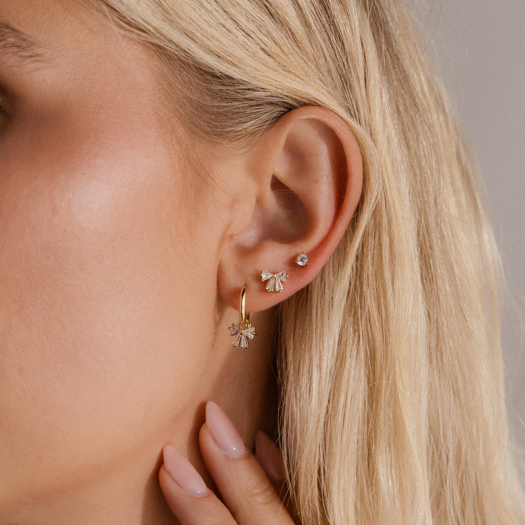 Close up of a woman wearing a gold Starlight Bow Hoop earring paired with matching bow stud and a small crystal stud, styled together for a chic layered ear look.