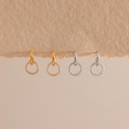 Two pairs of small hoop earrings with dangling open circles encrusted with clear stones, shown in gold and silver finishes, hanging against a textured beige surface.