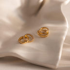 Pair of gold hoop earrings with interlocking circles, one smooth and one encrusted with small clear stones, displayed on a softly curved cream colored surface.