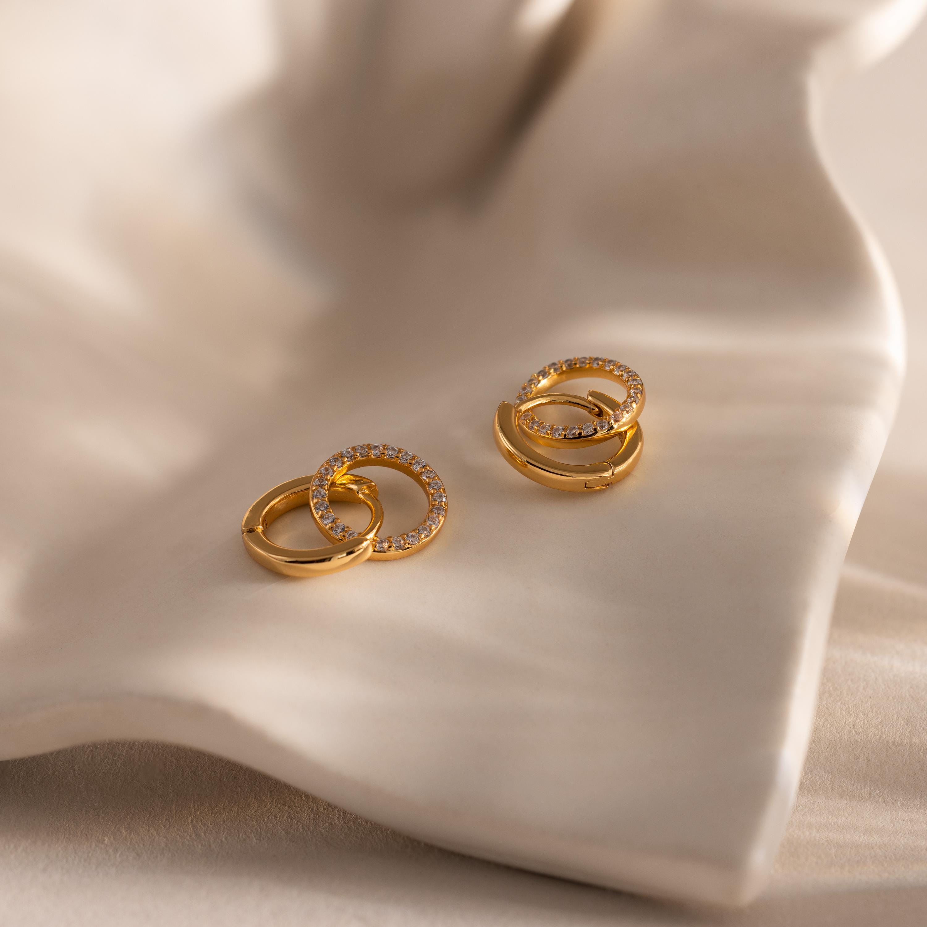 Pair of gold hoop earrings with interlocking circles, one smooth and one encrusted with small clear stones, displayed on a softly curved cream colored surface.
