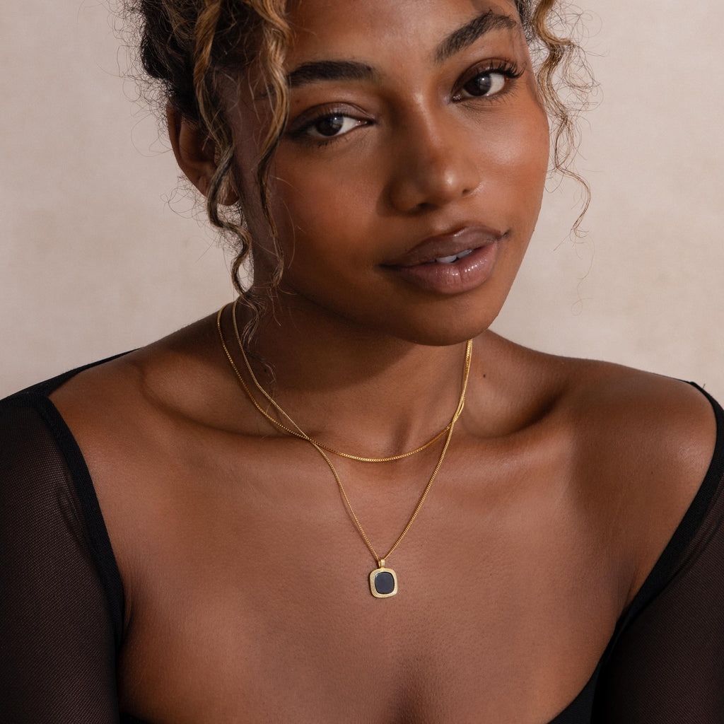A woman wearing the Square Onyx Necklace layered with gold necklaces over a sheer black top gazes at the camera with a soft expression—the necklace’s striking black onyx gemstone stands out as a bold statement piece.