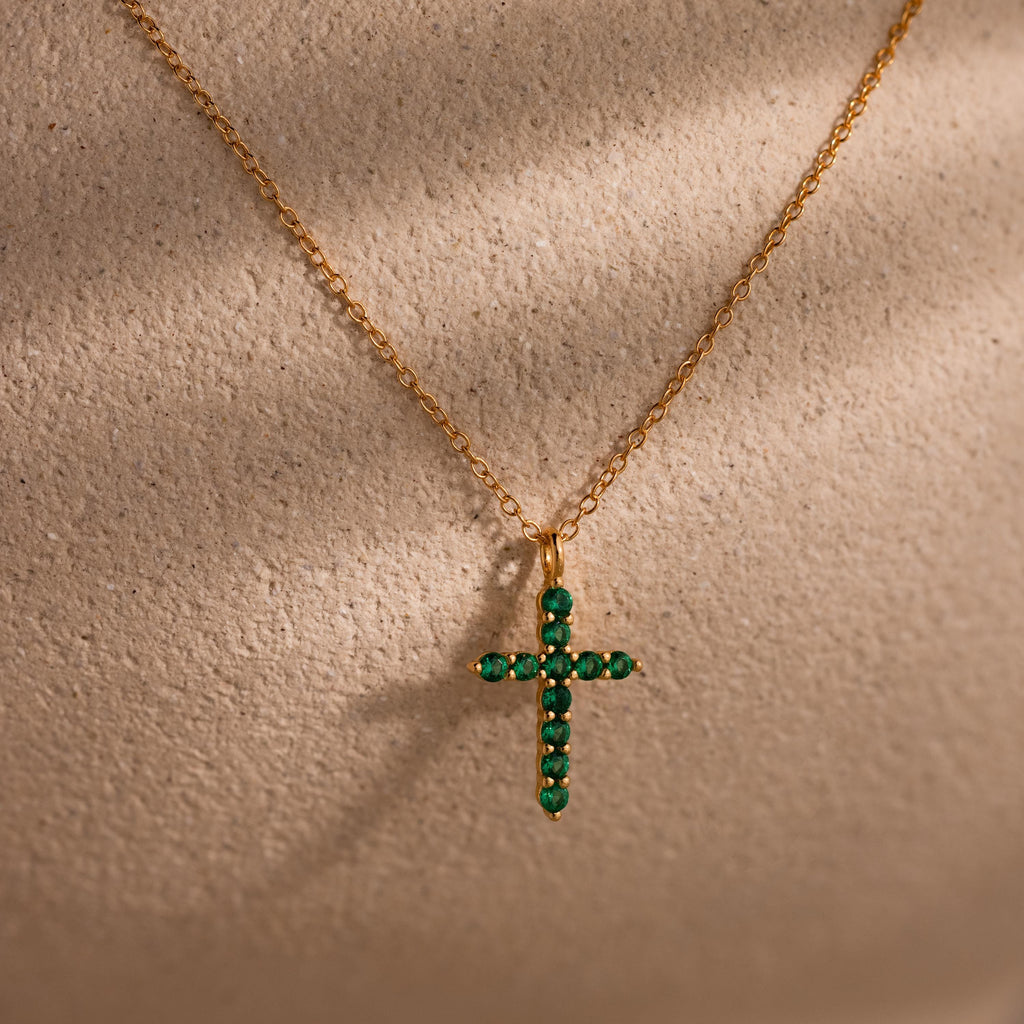 Gold chain necklace featuring a cross pendant adorned with small round green gemstones, displayed against a textured beige background.