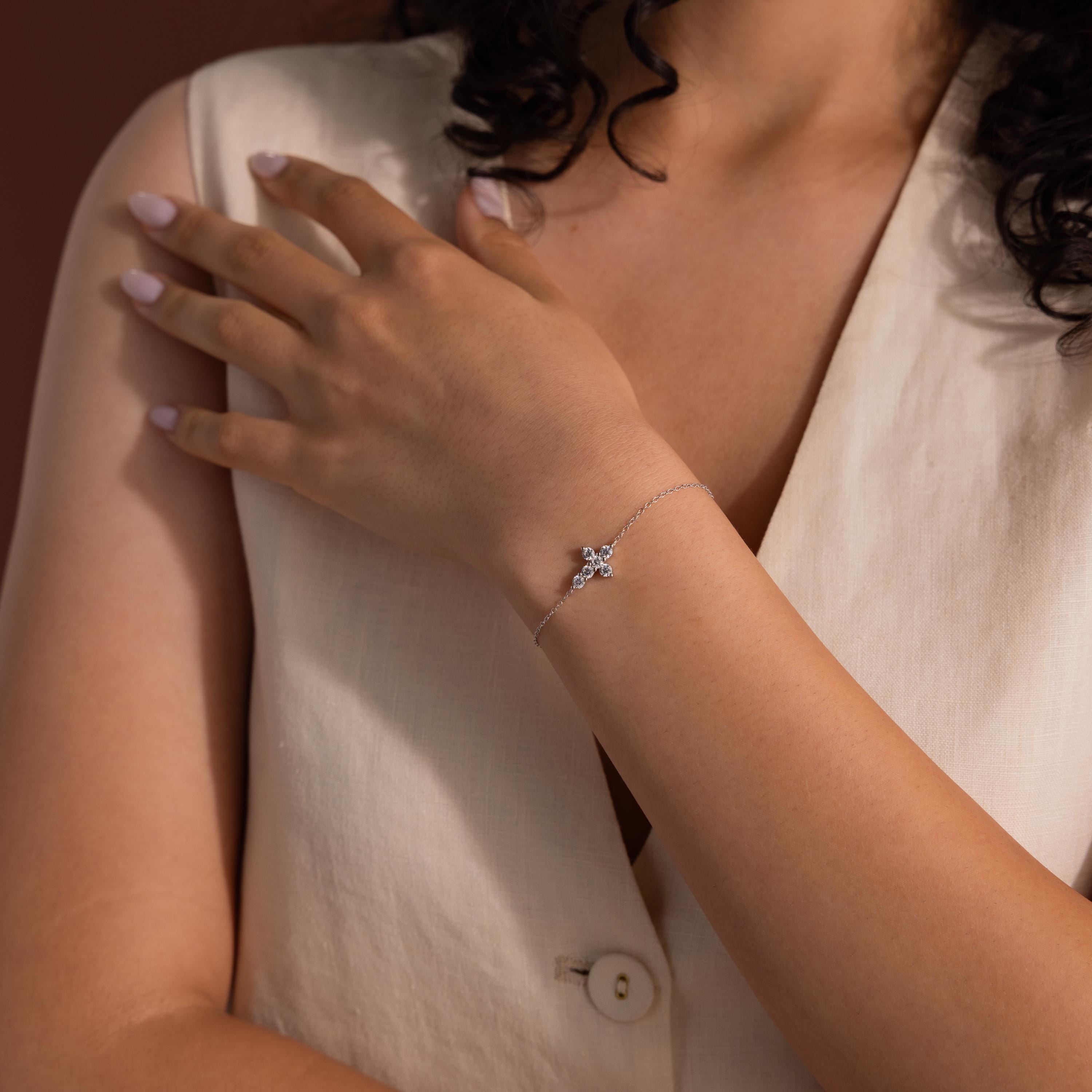 Woman wearing a delicate silver chain bracelet featuring a small cross-shaped charm adorned with round cut white gemstones, styled with a cream sleeveless top.