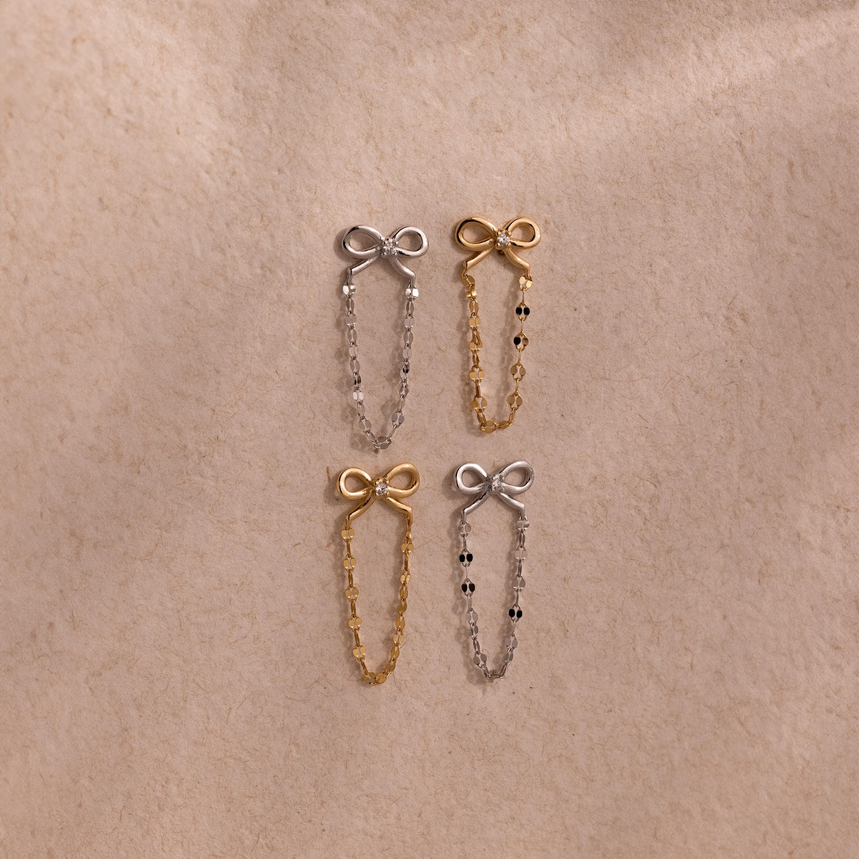 Four Bow Chain Studs in gold and silver feature delicate chain accents. Displayed on beige fabric, these feminine, vintage-inspired earrings are a charming jewelry addition.