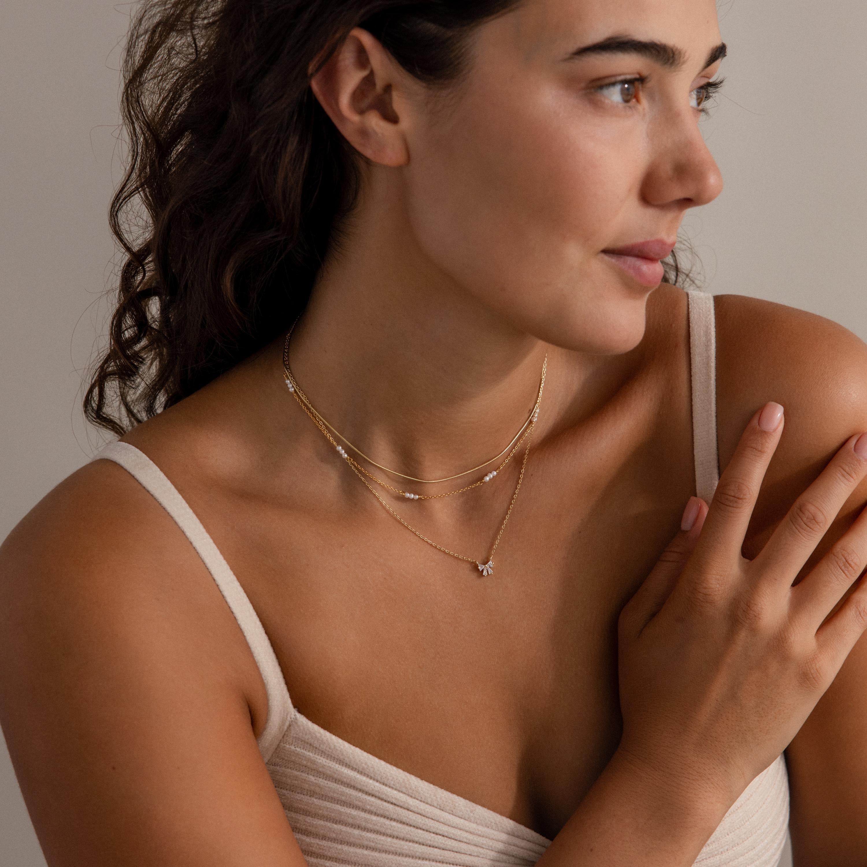 Woman with curly hair wearing layered gold plated necklaces, including the Diamond Bow Necklace featuring sparkling cubic zirconia, and a beige tank top, looking to the side.
