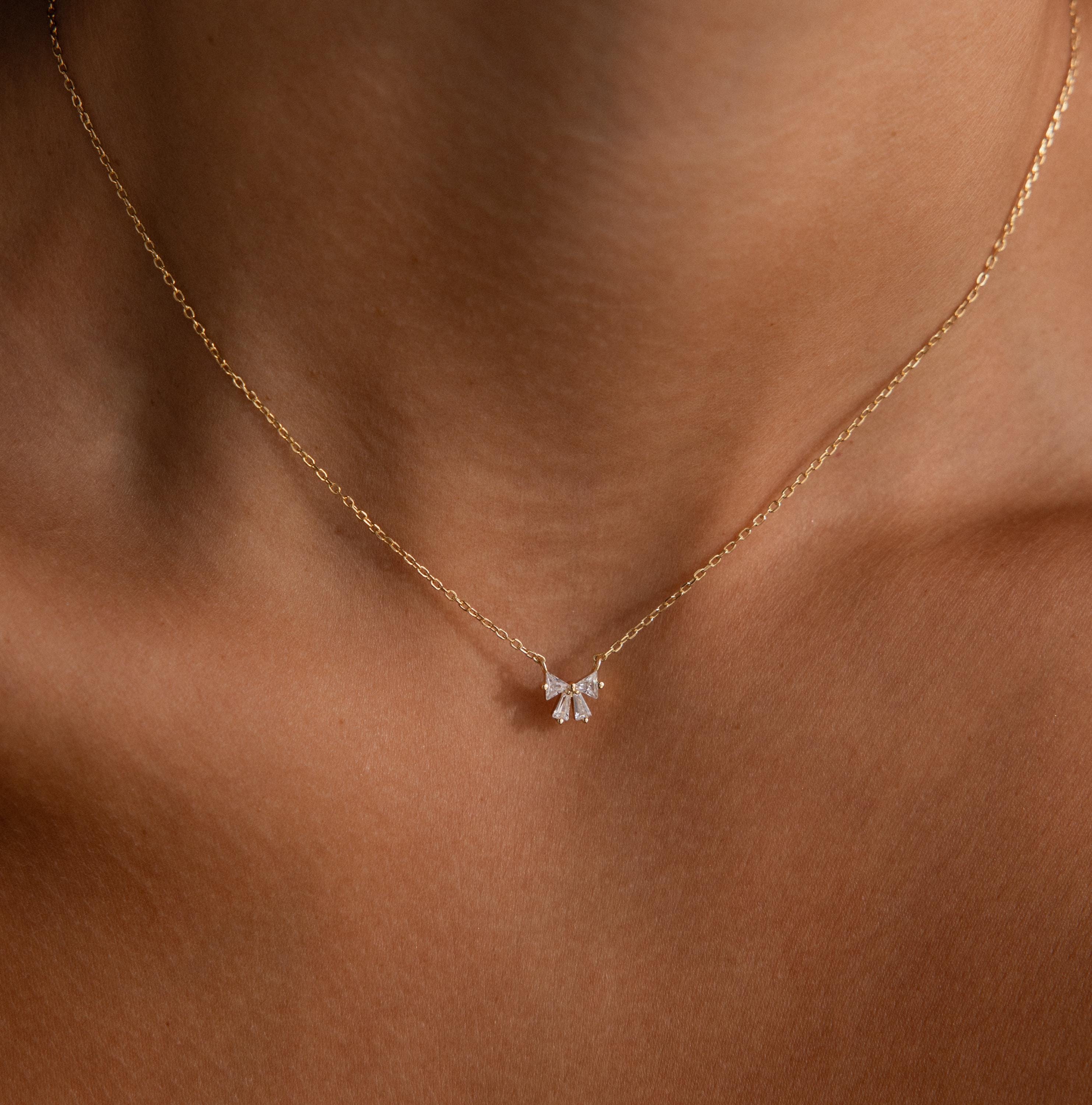 The Diamond Bow Necklace in 18K Gold features a graceful bow design adorned with sparkling stones, elegantly resting on the wearers neck.