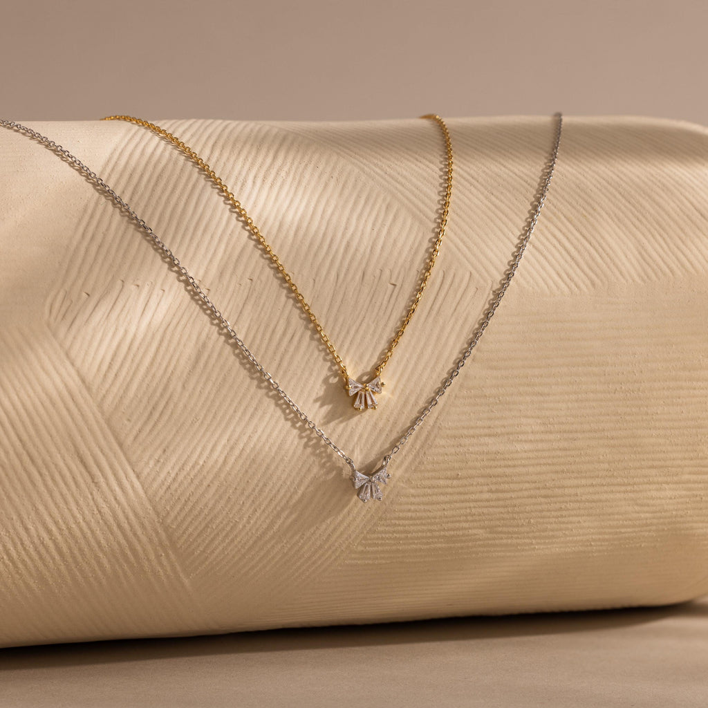 Two elegant Diamond Bow Necklaces, one gold plated and one silver, are displayed on a textured cream surface.