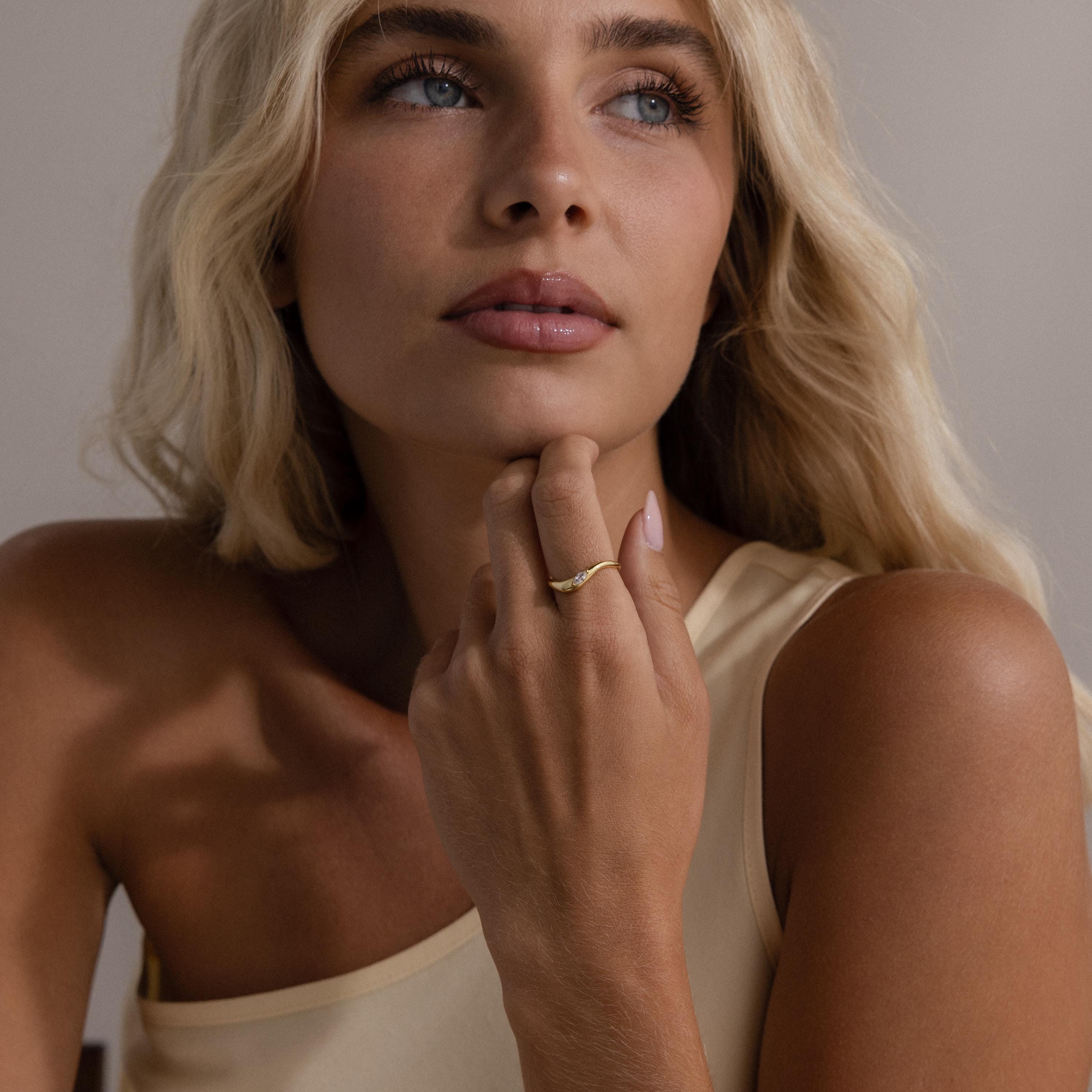 A woman with long blonde hair wears a one-shoulder beige top and gazes upward, her hand adorned with the Zen Diamond Curve Ring resting on her chin.