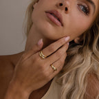 A woman with blonde hair touches her face, wearing the Zen Diamond Curve Ring and other gold rings. She has natural makeup and a neutral expression.