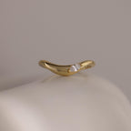 The Zen Diamond Curve Ring features a single round marquise diamond accent in a subtle wave design, crafted in gold and displayed on a smooth, off-white surface.