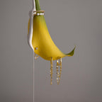 A yellow flower petal adorned with pearls and Duo Mirror Chain Huggies in 18K Gold hangs elegantly against a plain gray background.