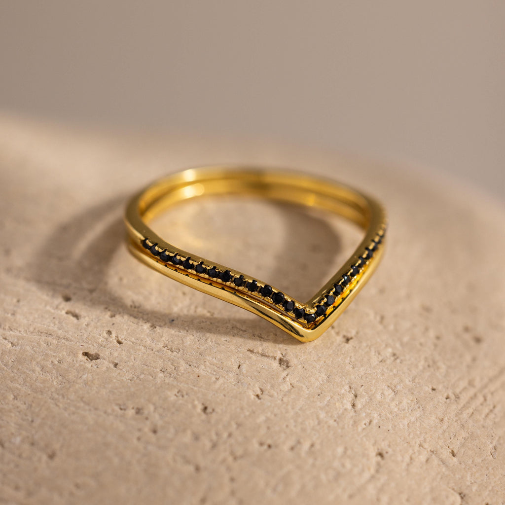 Close-up of a gold chevron ring with a row of pavé-set black stones, displayed on a textured neutral surface.