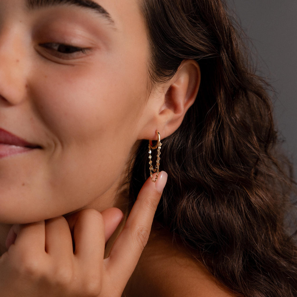 A woman with wavy brown hair smiles, touching a dangling earring from her Duo Mirror Chain Huggies in 18K Gold—an elegant accent to her modern layered jewelry look.