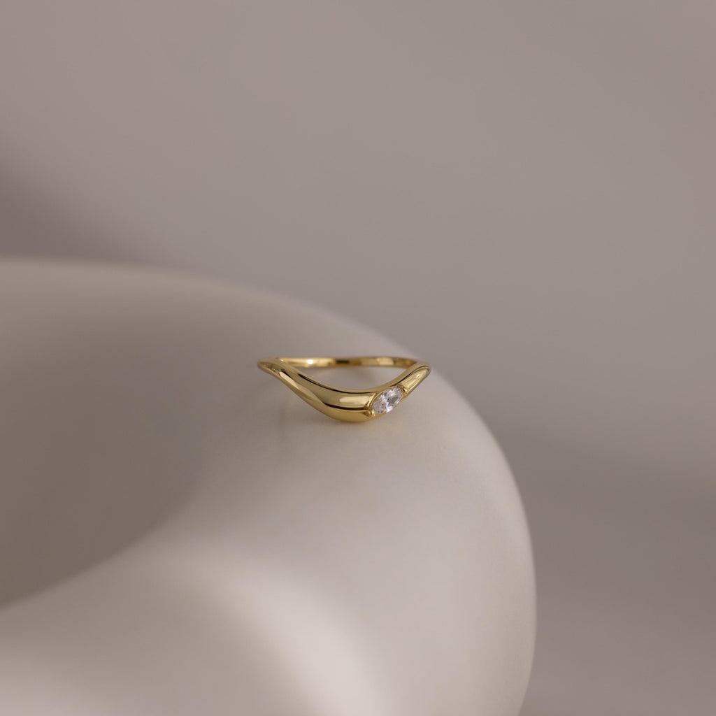 The Zen Diamond Curve Ring is a minimalist piece with a sleek open design and a single marquise diamond accent, beautifully presented on a smooth, light-colored surface.