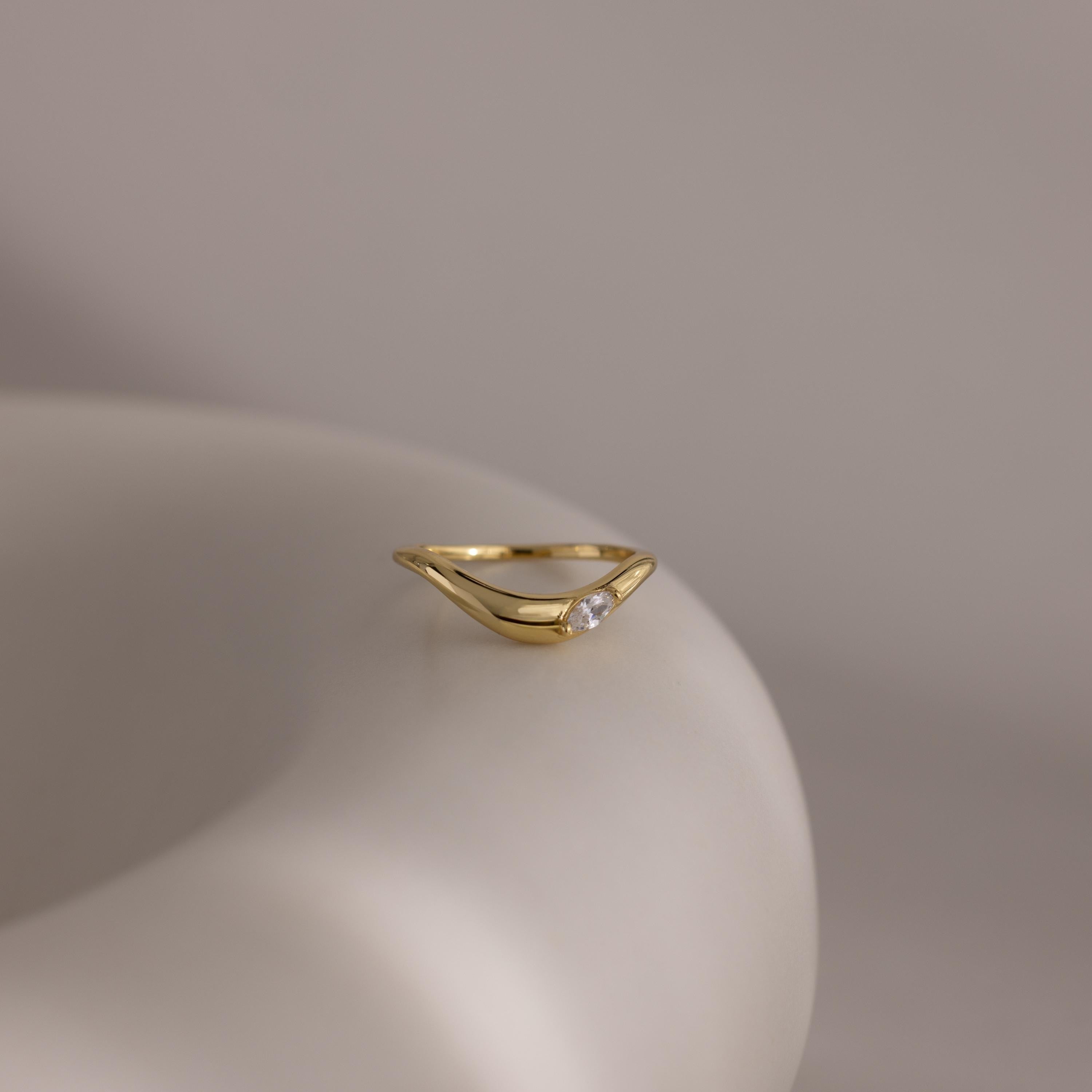 The Zen Diamond Curve Ring is a minimalist piece with a sleek open design and a single marquise diamond accent, beautifully presented on a smooth, light-colored surface.