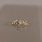 The Diamond Curve Cluster Ring in 18K Gold, adorned with a Baguette Stone in the center and a cluster of marquise and round diamonds on the sides, is shown on a reflective surface.
