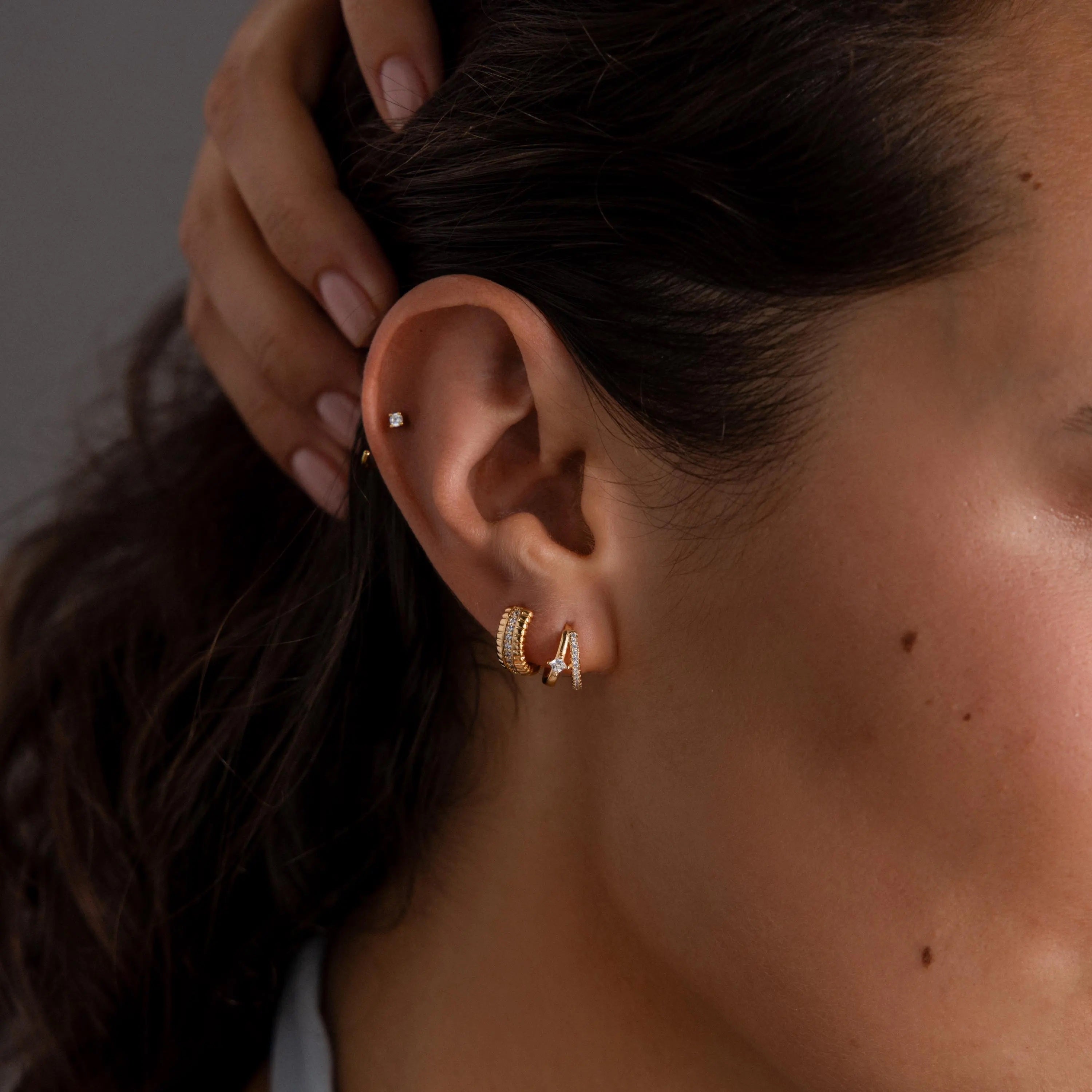 Close-up of a woman's ear with three elegant earrings, featuring the Pave Diamond Duo Huggies in 18K Gold and a small stud, as she gently touches her hair.