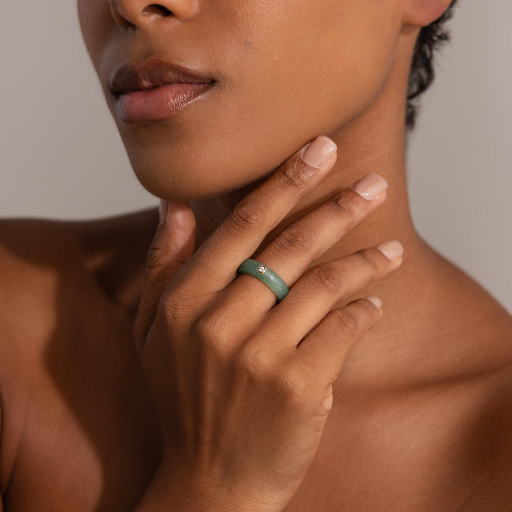 A woman with natural nails touches her face, wearing the Jade Diamond Ring—a green jade stone set in gold and accented with a small diamond.