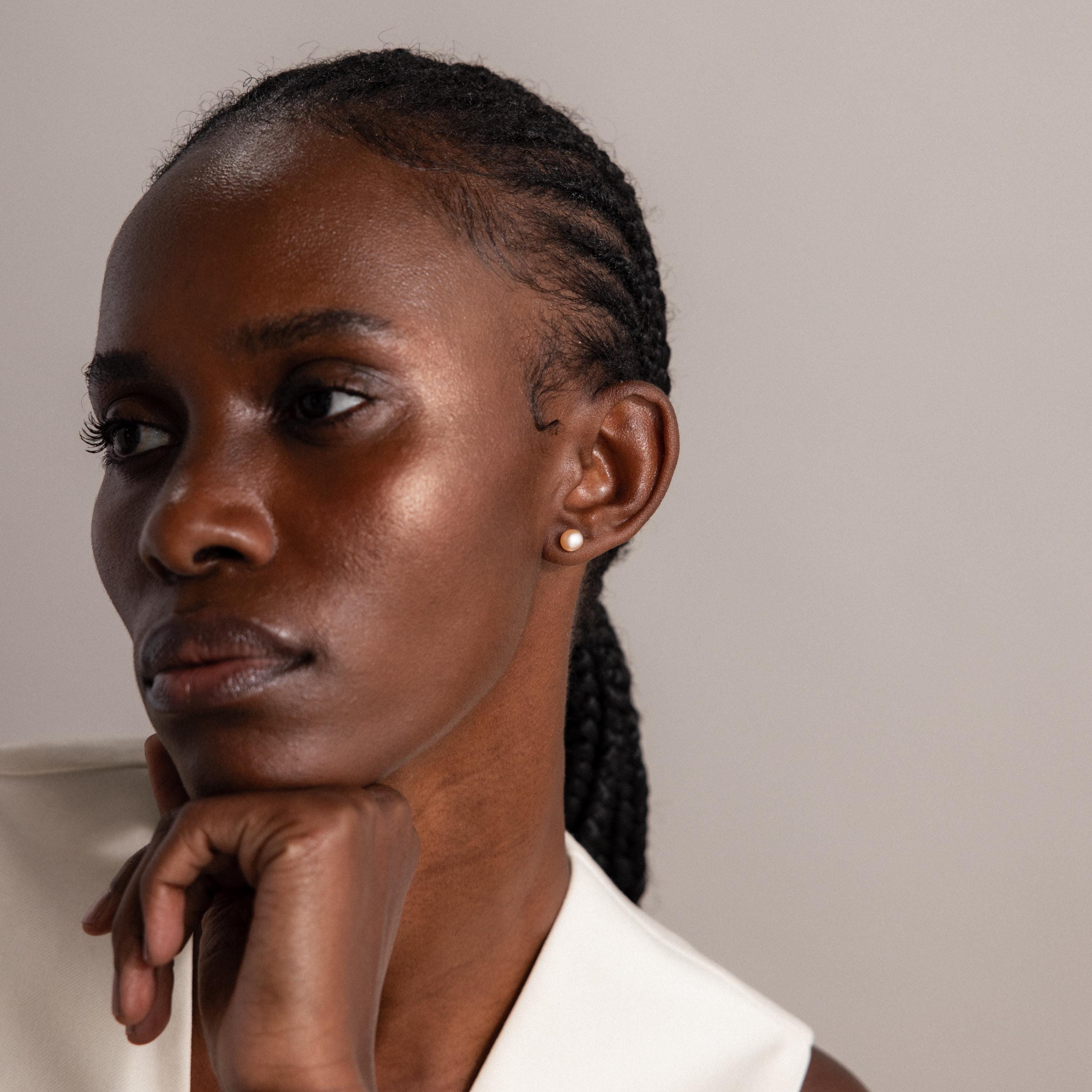 A woman with braided hair, wearing Pink Pearl Studs earrings, rests her chin on her hand and gazes thoughtfully to the side.