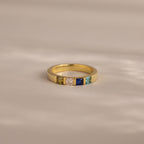 The Multiple Birthstone Band features five colorful square gemstones set in a row on a soft beige surface.