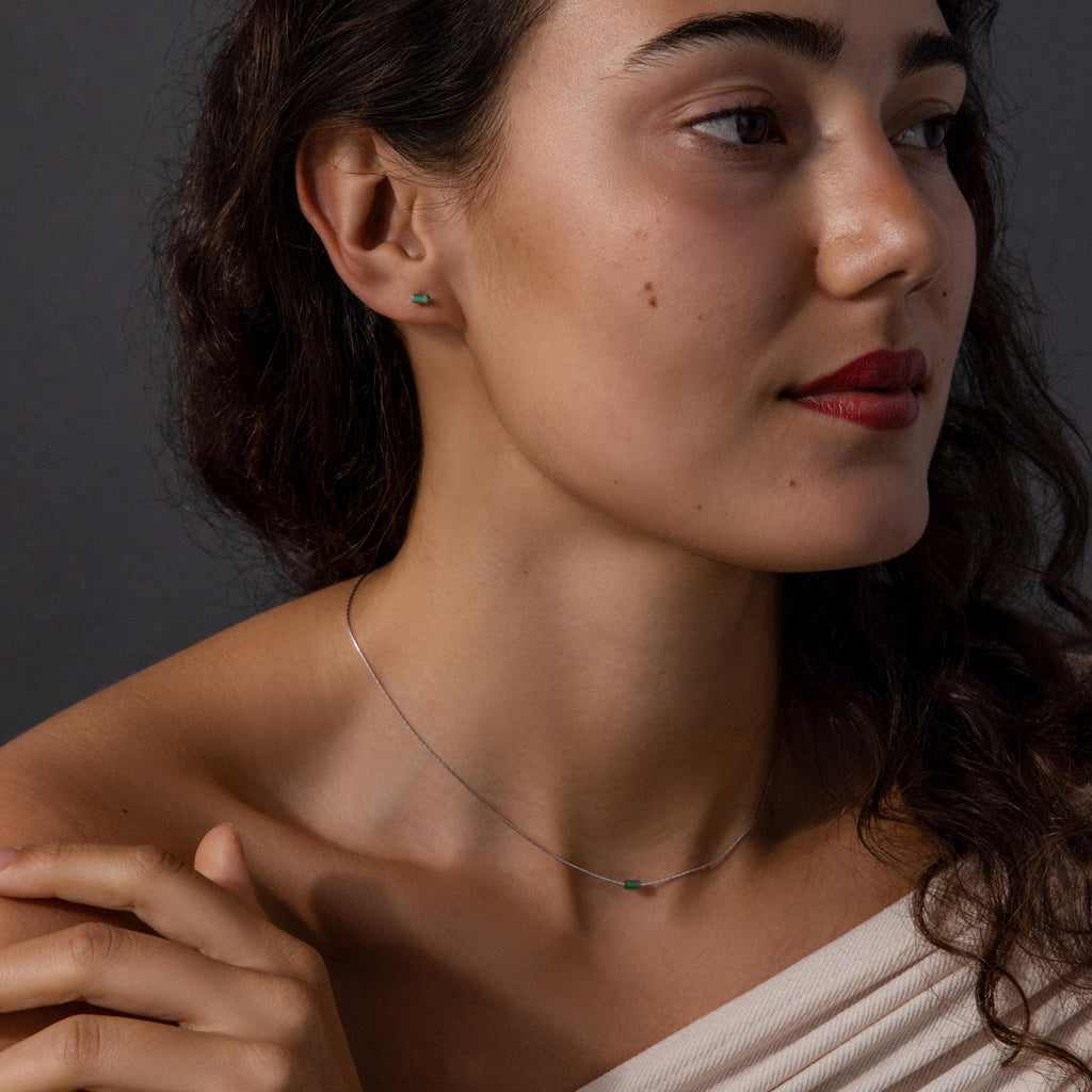 Woman with wavy hair wears the Jade Bar Jewelry Set in Sterling Silver, featuring a silver necklace and small green stud earrings, as she looks to the side.