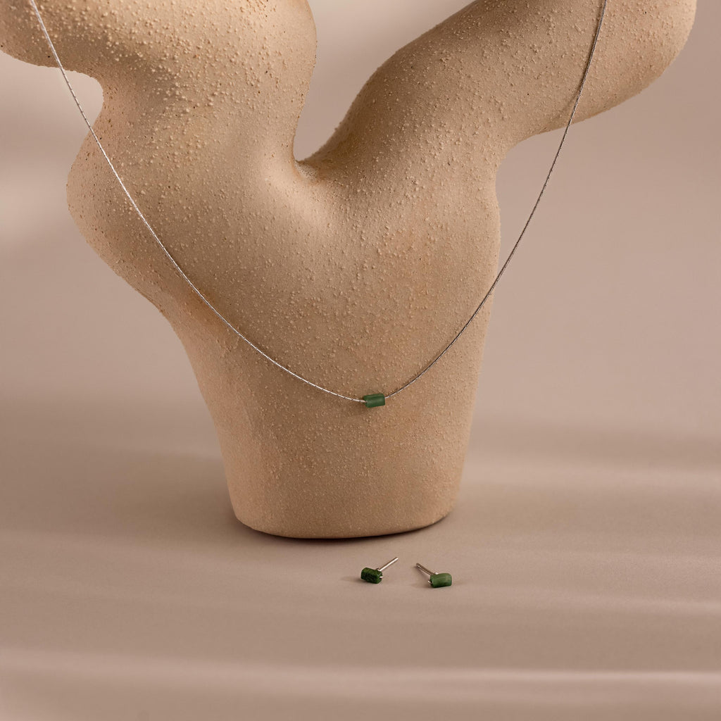 The Jade Bar Jewelry Set in Sterling Silver, featuring a bar necklace and stud earrings with small green jade stones, is displayed on a beige textured abstract stand.