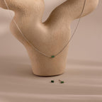 The Jade Bar Jewelry Set in Sterling Silver, featuring a bar necklace and stud earrings with small green jade stones, is displayed on a beige textured abstract stand.