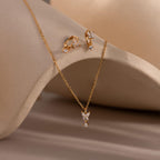 The Diamond Butterfly Drop Jewelry Set, featuring sparkling huggie earrings and a butterfly drop necklace, is elegantly displayed on a neutral curved surface.