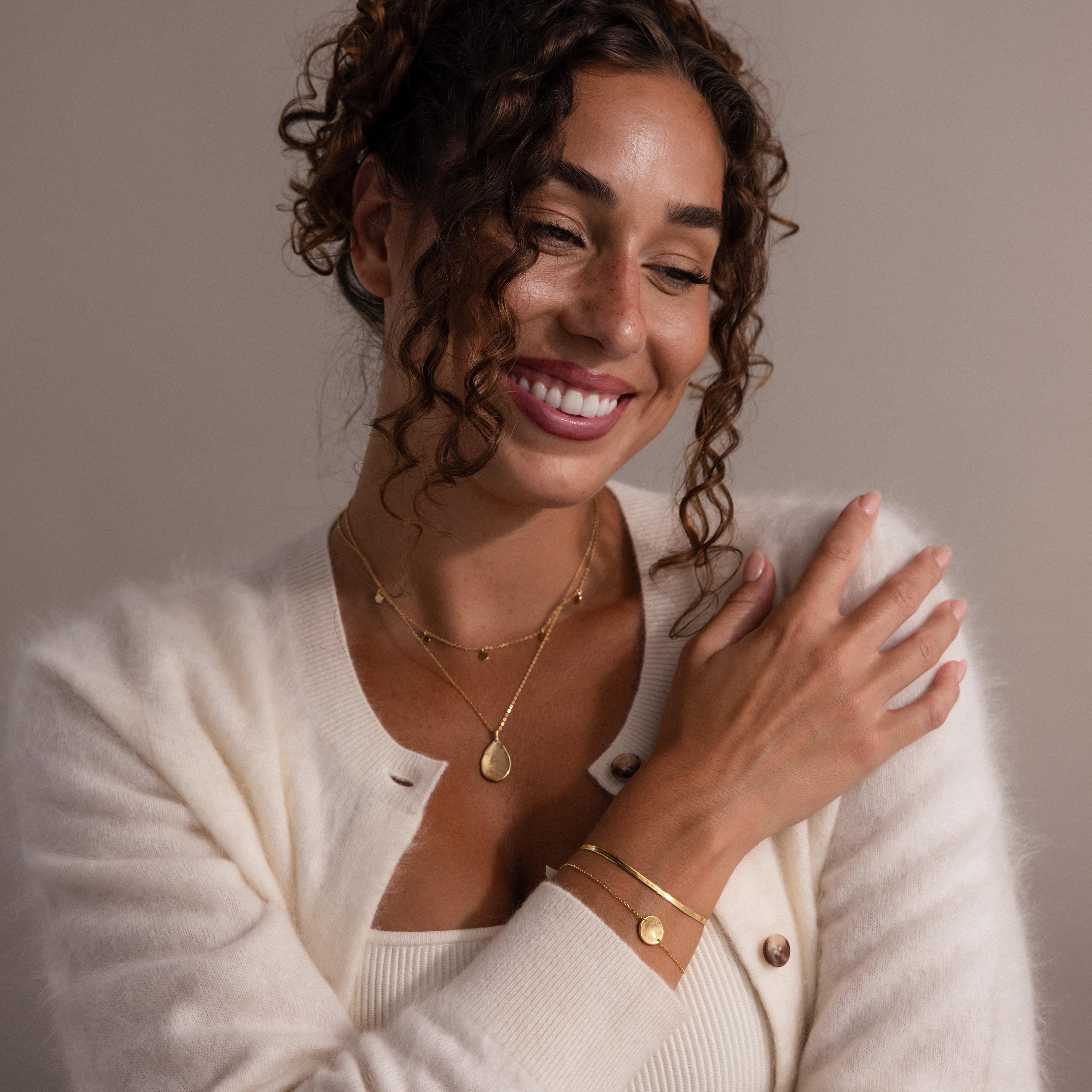 Smiling woman with curly hair wears a cream cardigan, ribbed top, and the Dainty Organic Fingerprint Bracelet in 18K Gold against a neutral background.