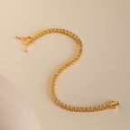 The Diamond Bezel Tennis Necklace & Bracelet Set in 18K Gold is displayed on a beige surface in an elegant S shape, showcasing its round diamonds.