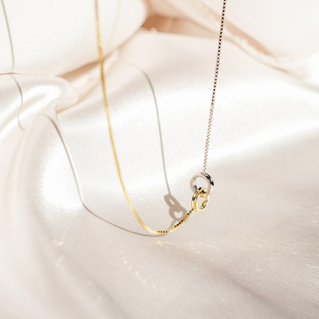 The Interlocking Circles Necklace and Bracelet Set, featuring gold and silver tones, rests on cream satin fabric, softly illuminated by natural light.