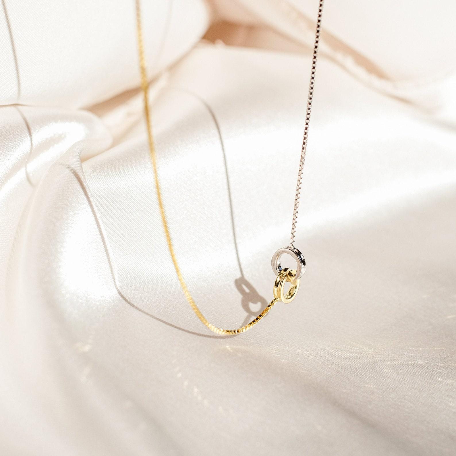The Interlocking Circles Necklace and Bracelet Set, featuring gold and silver tones, rests on cream satin fabric, softly illuminated by natural light.