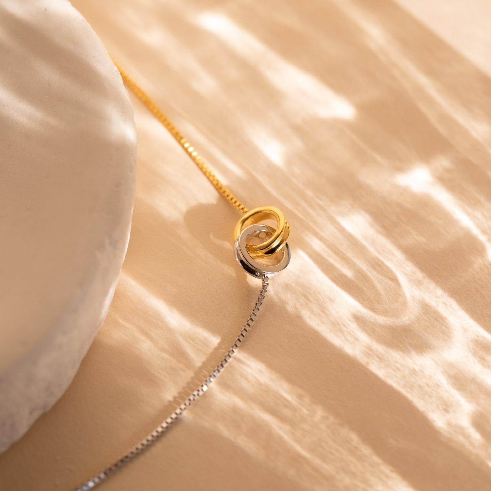 The Interlocking Circles Necklace and Bracelet Set features gold and silver interlocked rings displayed on a beige surface with soft, wavy light reflections—a timeless addition to our jewelry collection.