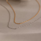 The Dainty Paperclip Necklace features delicate rectangular links in gold and silver, capturing timeless style—perfect for effortless layering and beautifully displayed on a soft beige surface.