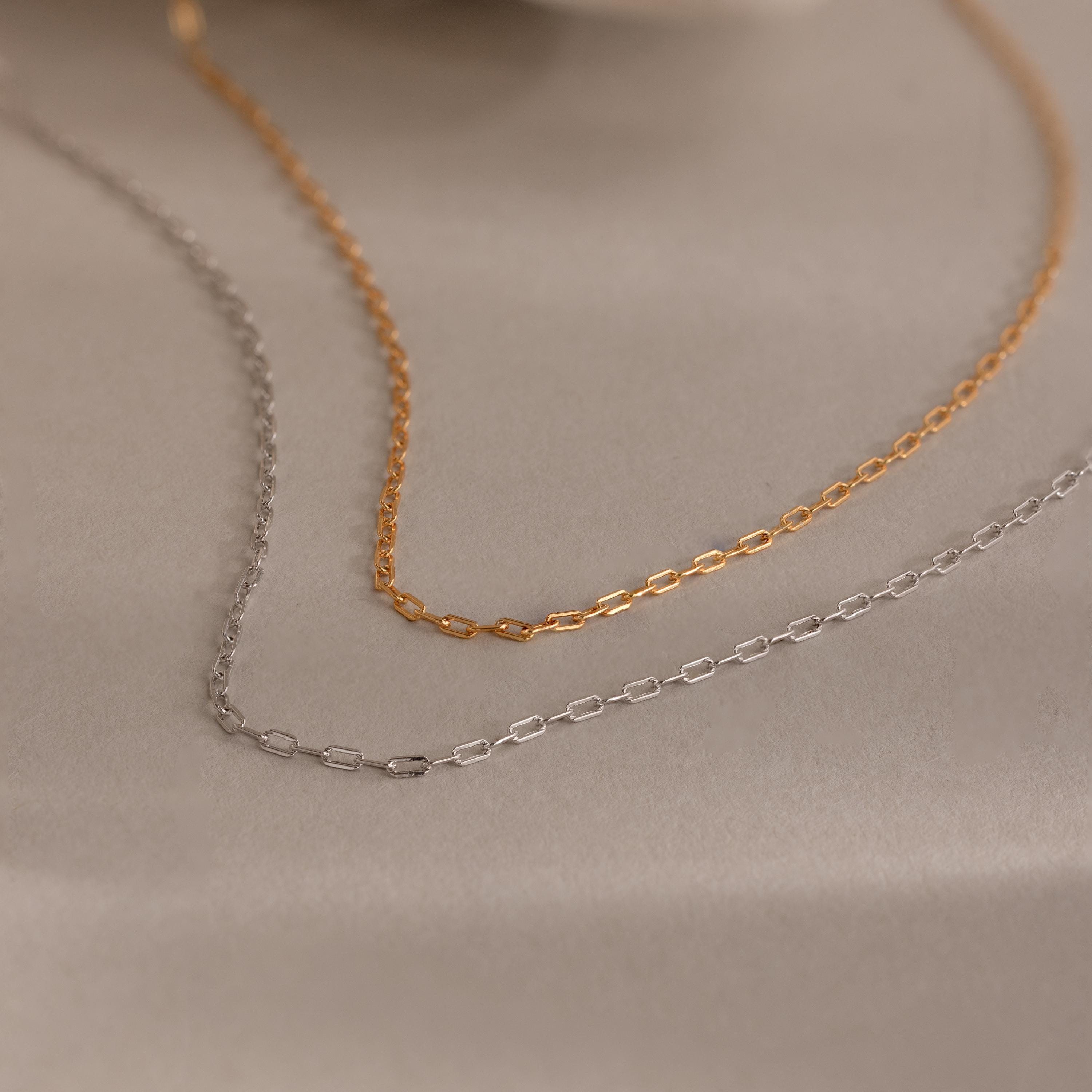 The Dainty Paperclip Necklace features delicate rectangular links in gold and silver, capturing timeless style—perfect for effortless layering and beautifully displayed on a soft beige surface.