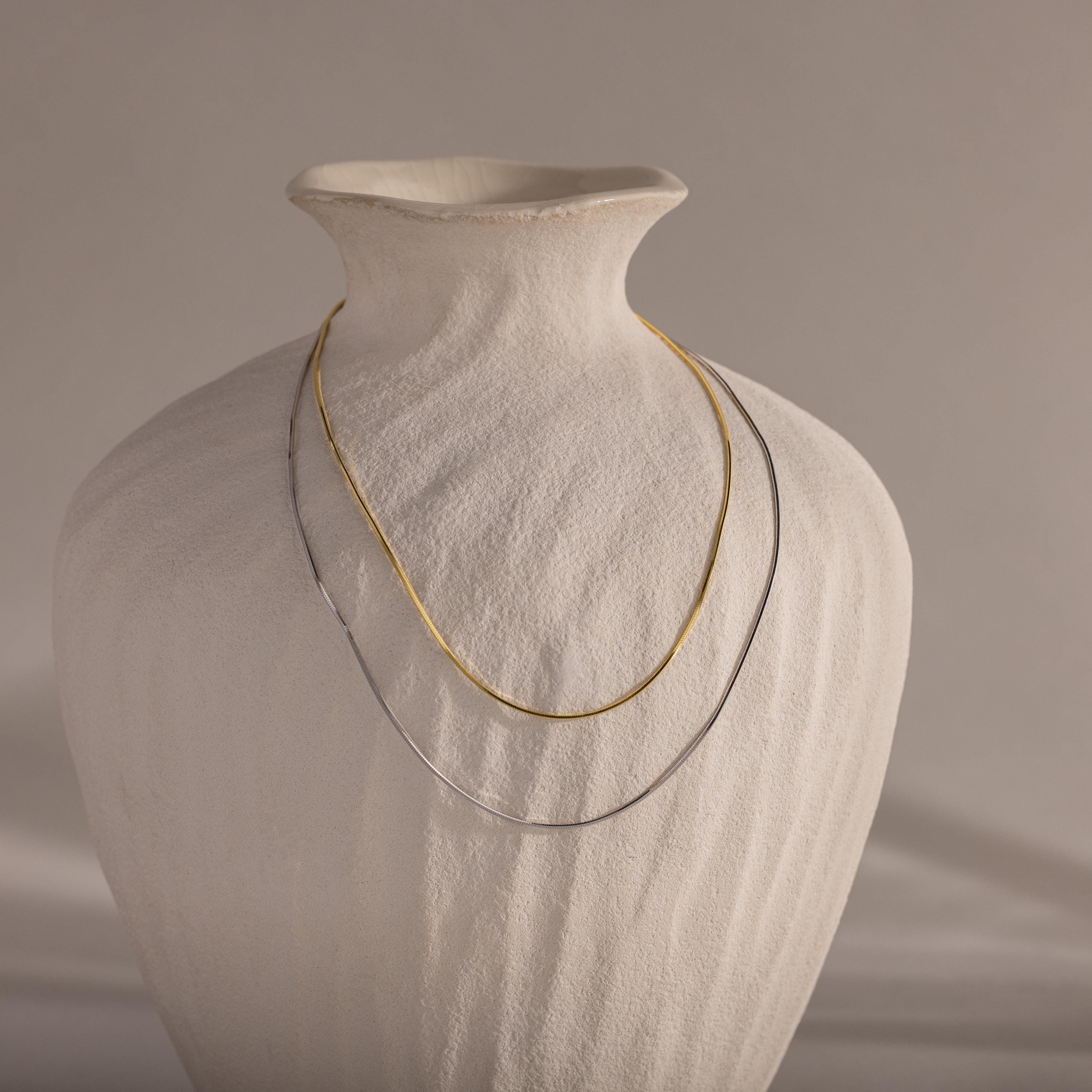 A Dainty Herringbone Necklace draped gracefully over a textured white ceramic vase.