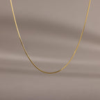 The Dainty Herringbone Necklace features a delicate curve, beautifully displayed against a simple beige background.