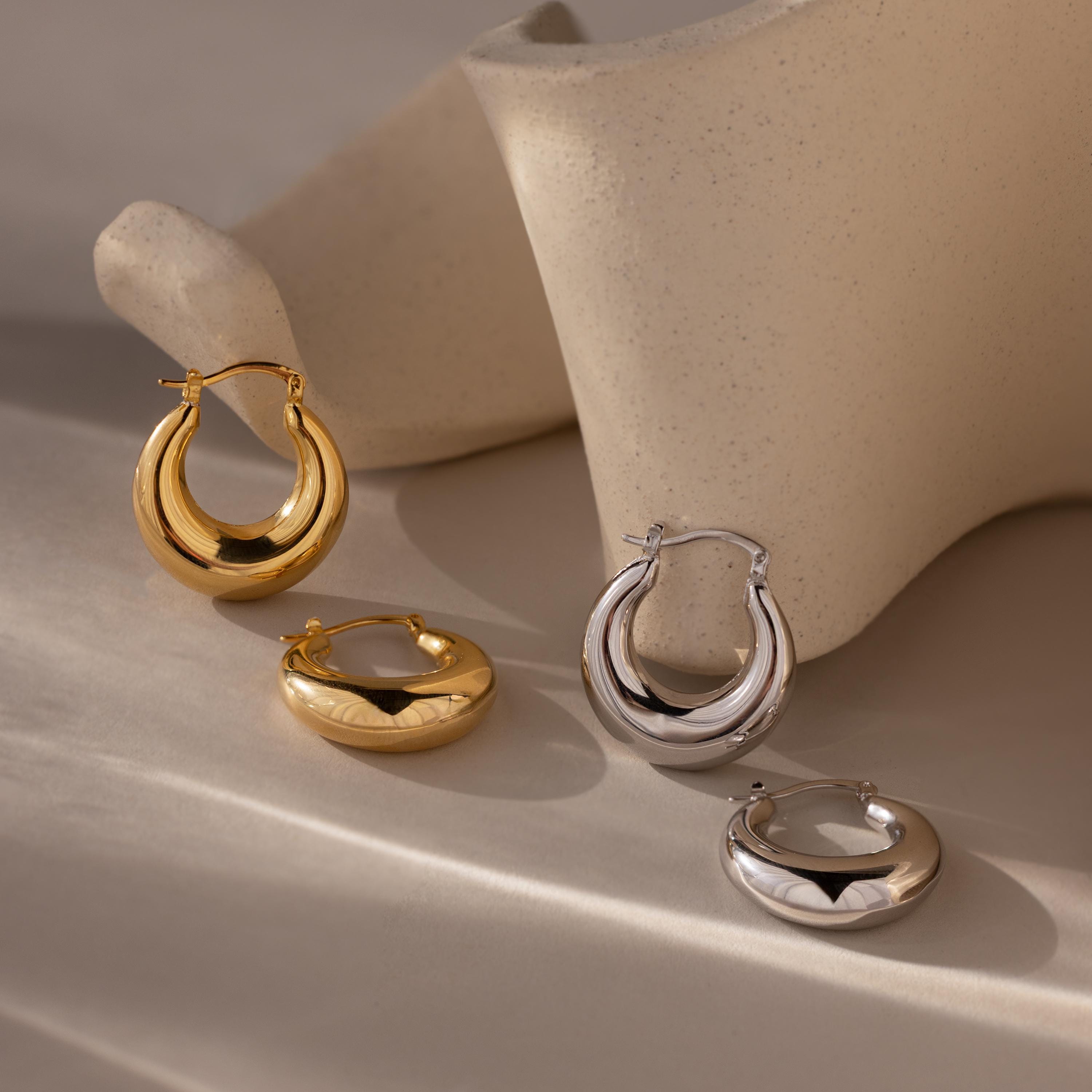 Sculpted Hoops in gold and silver, featuring a high-shine finish, are displayed on a soft beige surface with abstract ceramic décor.